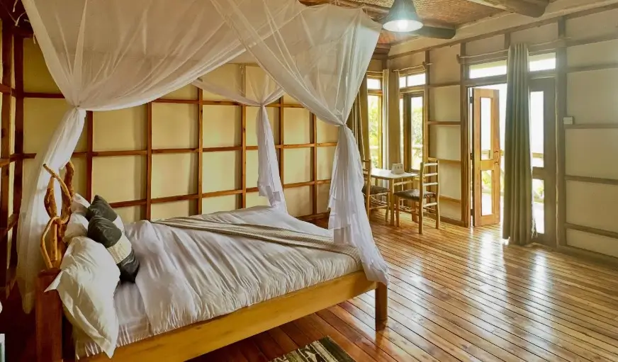 A bright and airy luxury bedroom with a four-poster bed, mosquito netting, and large windows facing the Bwindi rainforest.