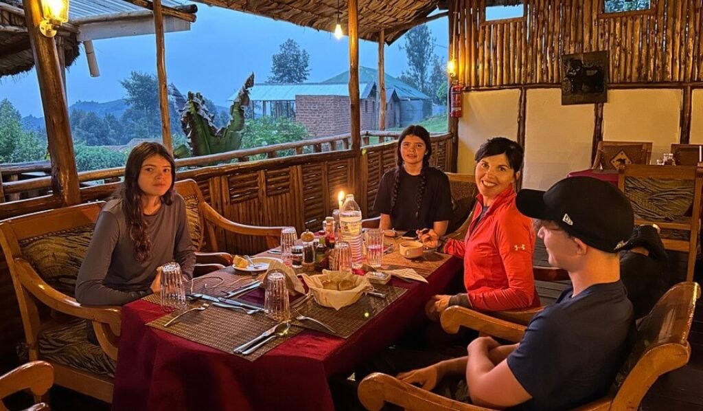 A group of travelers enjoying a meal at a long table on an outdoor balcony with panoramic views of the Ruhija hills.