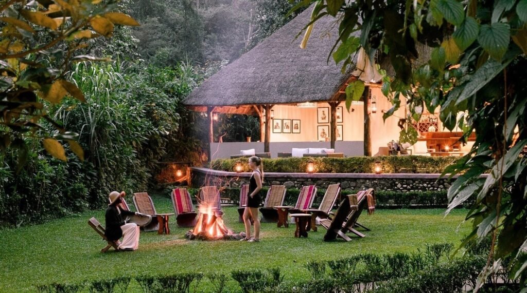 A stone and thatch luxury lodge nestled in the forest with a communal campfire area lit at dusk.