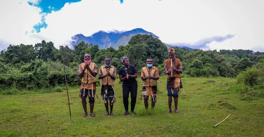 A group of Batwa men and women performing a traditional cultural dance in a lush green clearing of Mgahinga Gorilla National Park, with a volcanic peak visible in the background.