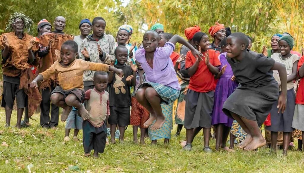 A large group of Batwa community members, including children and adults, performing a traditional dance in a grassy forest clearing.