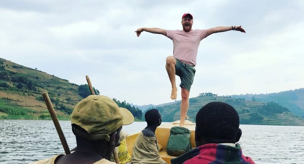 A traveler balancing on the edge of a wooden boat with arms outstretched, celebrating the journey with the rolling hills of the region in the background.