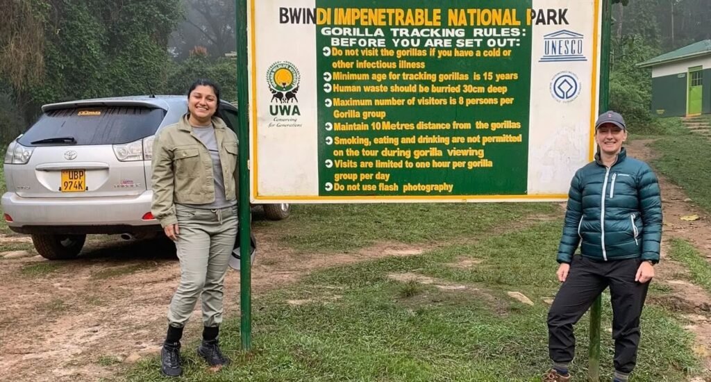 Two trekkers standing in front of the official wooden entrance sign for Bwindi Impenetrable National Park, marking the start of the trekking adventure.