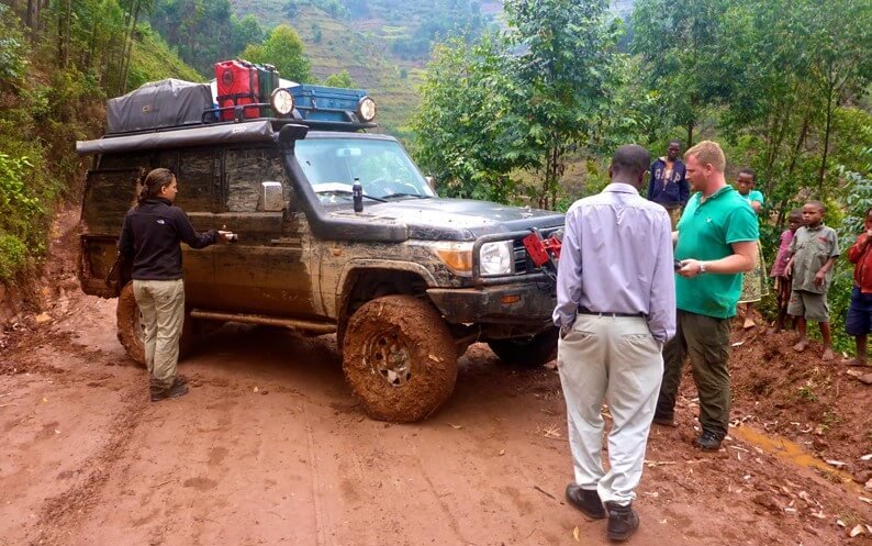 4WD safari vehicle on a muddy approach road to Bwindi during wet season, with trekkers nearby