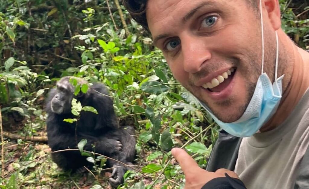 A male traveler wearing a face mask smiles beside a chimpanzee during a Kibale trekking experience