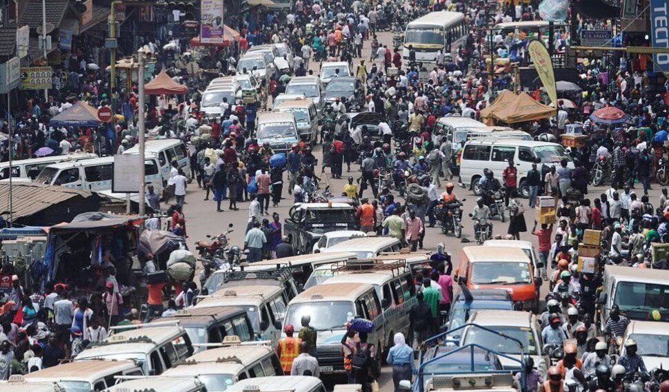 An aerial, high-angle view of a bustling street in downtown Kampala filled with white commuter taxis and a dense crowd of people, illustrating the organized chaos of the city’s commercial heart.