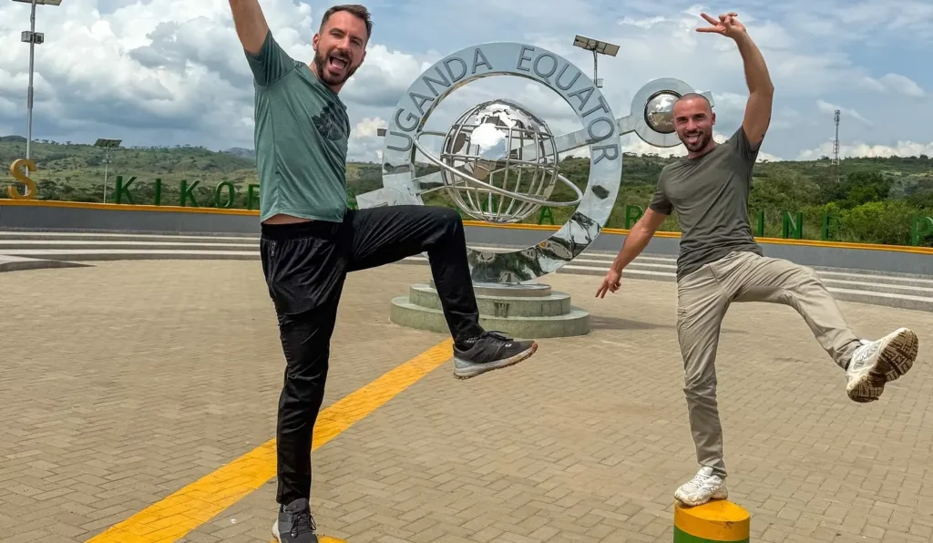 Two travelers pose playfully at the Uganda Equator landmark, standing near the circular metal globe monument that marks the crossing of the northern and southern hemispheres.