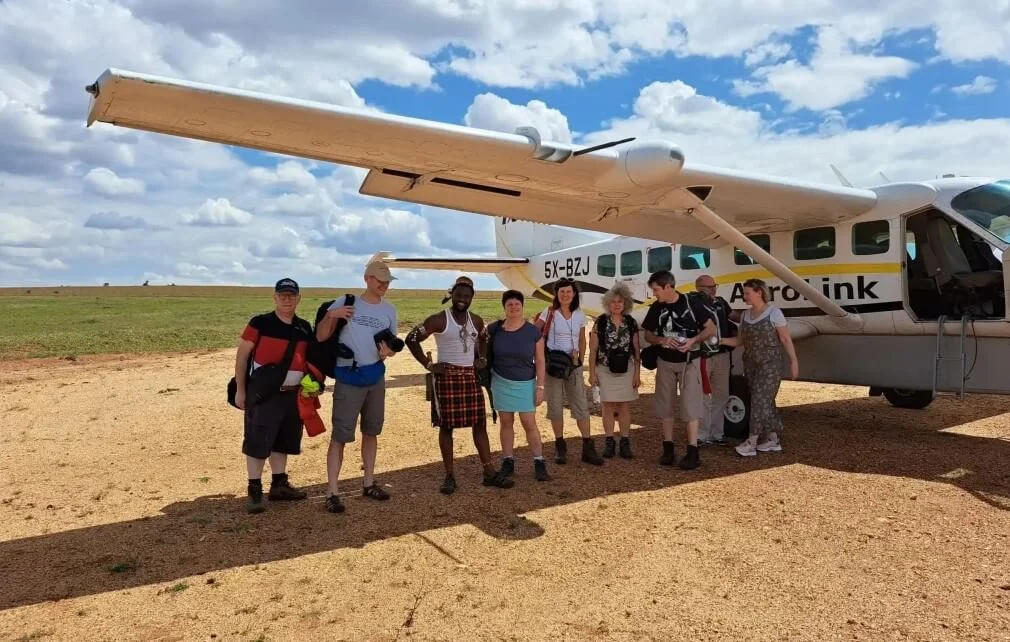 A group of travelers stands on a dirt airstrip in rural Uganda next to a small white AeroLink bush plane used for domestic flights to Bwindi.