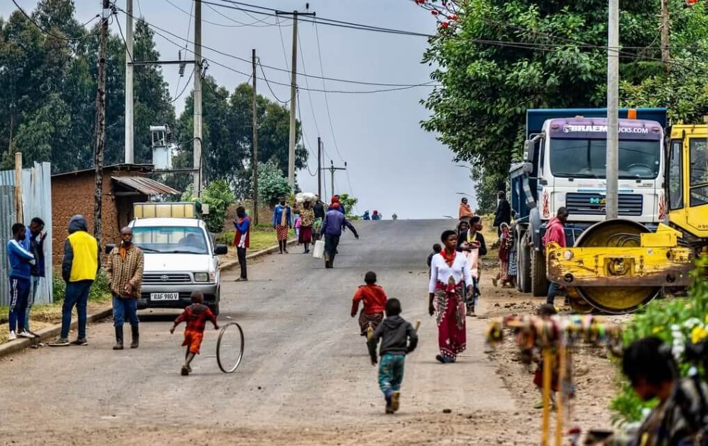 A paved road leading through the Goli border crossing between Uganda and the Democratic Republic of Congo, featuring local pedestrians, small shops, and a clear sky.