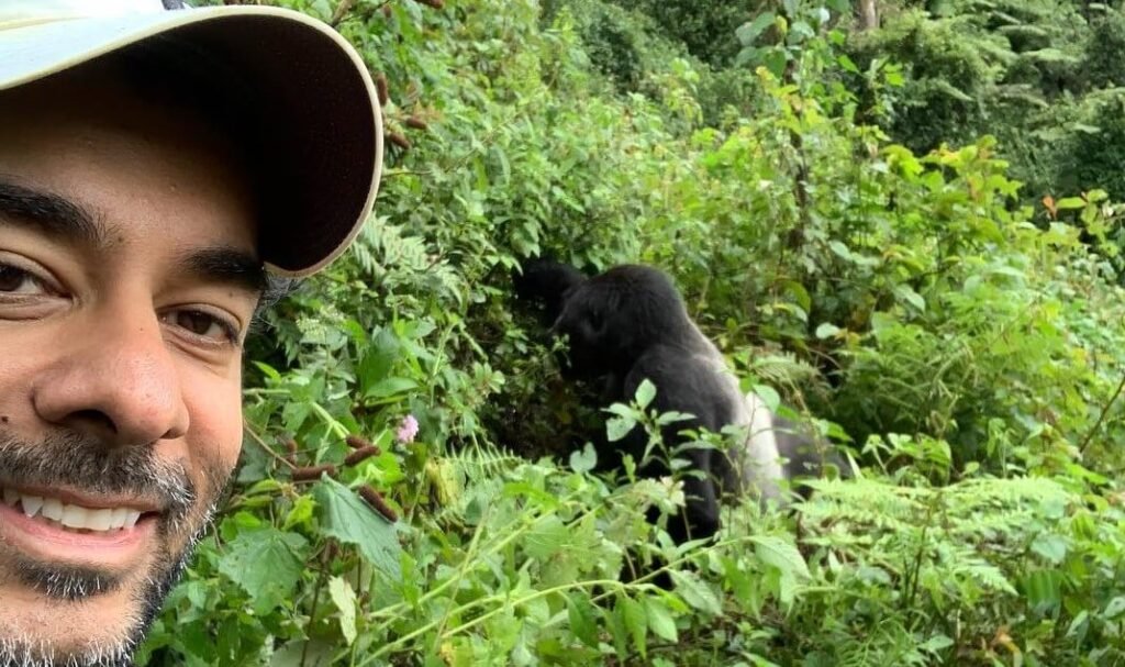 smiling man taking a careful selfie with a mountain gorilla visible in the background greenery.