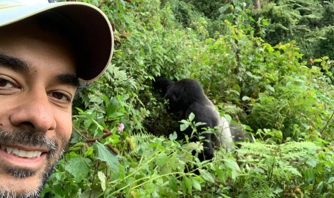 smiling man taking a careful selfie with a mountain gorilla visible in the background greenery.