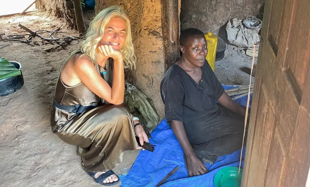 A female traveler sitting cross-legged on the ground inside a traditional Ugandan home, engaging in a quiet conversation with a local woman seated on a blue mat.