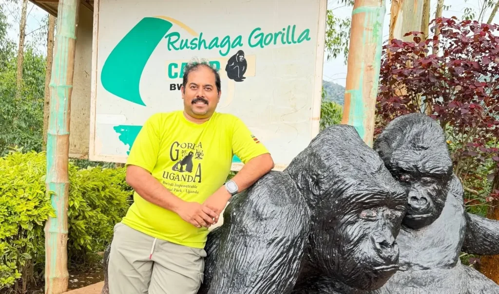 A man in a yellow shirt leans against a large black gorilla statue in front of the Rushaga Gorilla Camp sign, marking the entrance to the southern sector of Bwindi Impenetrable National Park.
