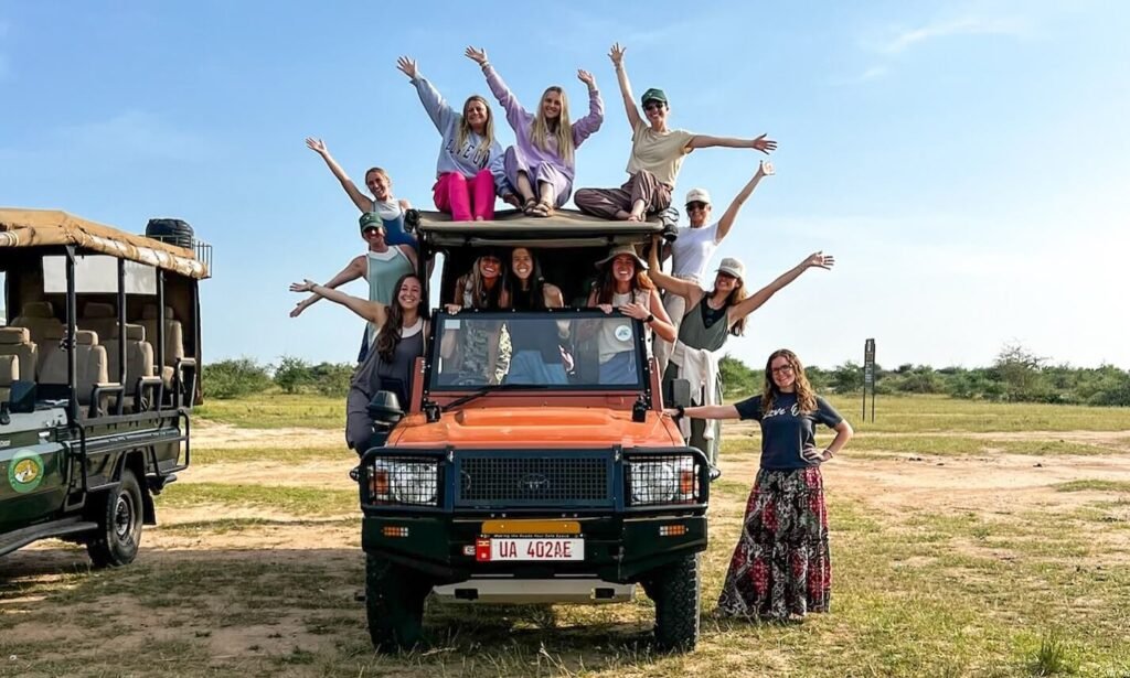 A group of smiling tourists posing on and around an open-top orange safari vehicle in a sunny Ugandan savanna, showcasing the social and adventurous nature of group travel.