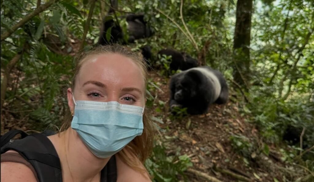A hiker wearing a blue surgical mask for conservation safety, taking a selfie with a gorilla resting in the background foliage.