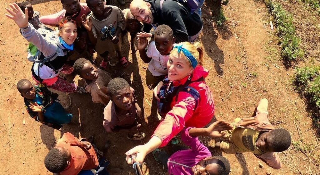 A group of local Ugandan children smiling and looking up at the camera while interacting with a traveler in a rural village.