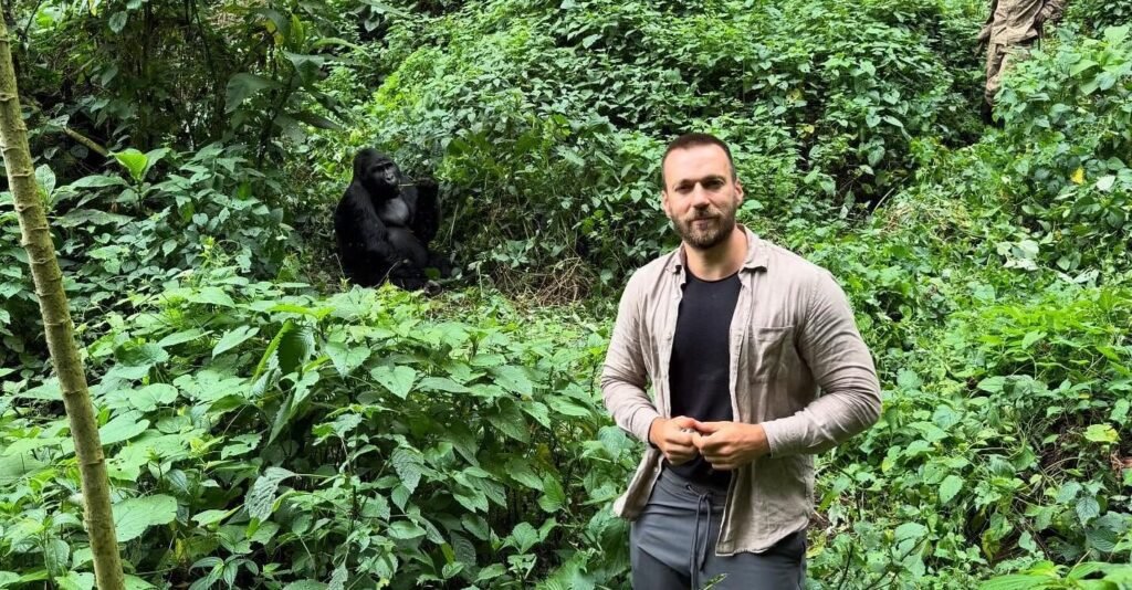 A male trekker standing on a narrow forest path with a silverback gorilla visible in the background, showing the proximity of the encounter.