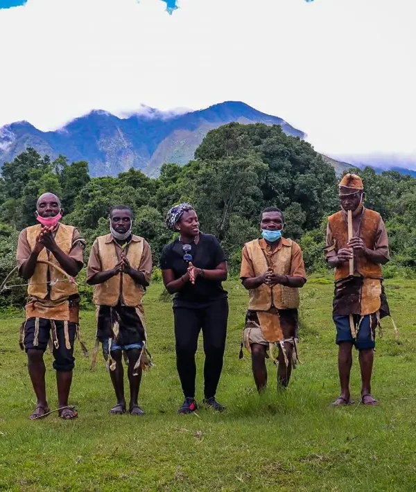 A group of Batwa men and women performing a traditional cultural dance in a lush green clearing of Mgahinga Gorilla National Park, with a volcanic peak visible in the background.