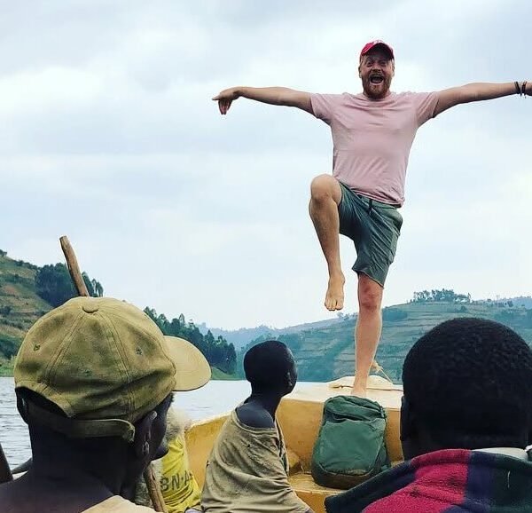 A traveler balancing on the edge of a wooden boat with arms outstretched, celebrating the journey with the rolling hills of the region in the background.