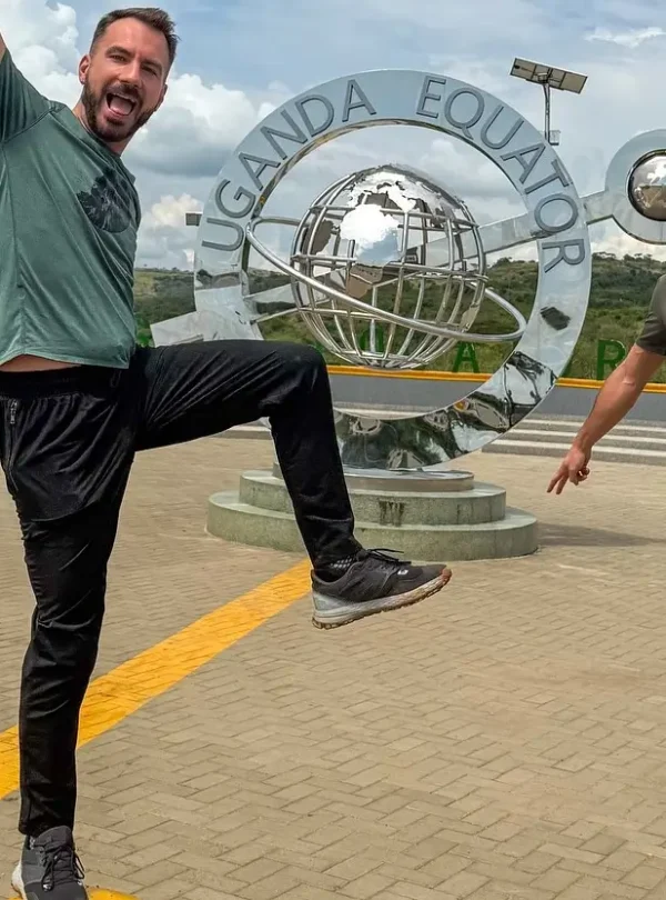 Two travelers pose playfully at the Uganda Equator landmark, standing near the circular metal globe monument that marks the crossing of the northern and southern hemispheres.
