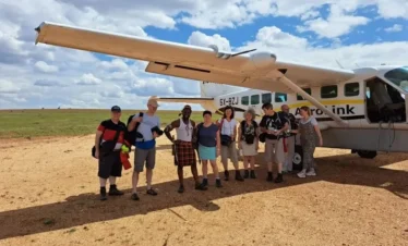 A group of travelers stands on a dirt airstrip in rural Uganda next to a small white AeroLink bush plane used for domestic flights to Bwindi.