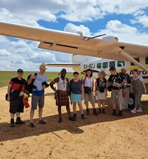 A group of travelers stands on a dirt airstrip in rural Uganda next to a small white AeroLink bush plane used for domestic flights to Bwindi.