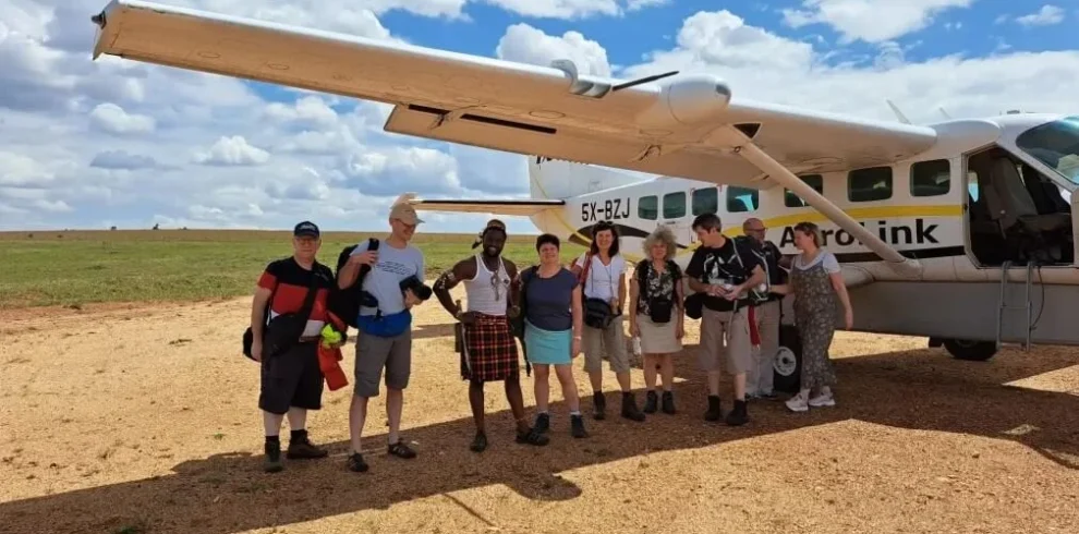 A group of travelers stands on a dirt airstrip in rural Uganda next to a small white AeroLink bush plane used for domestic flights to Bwindi.