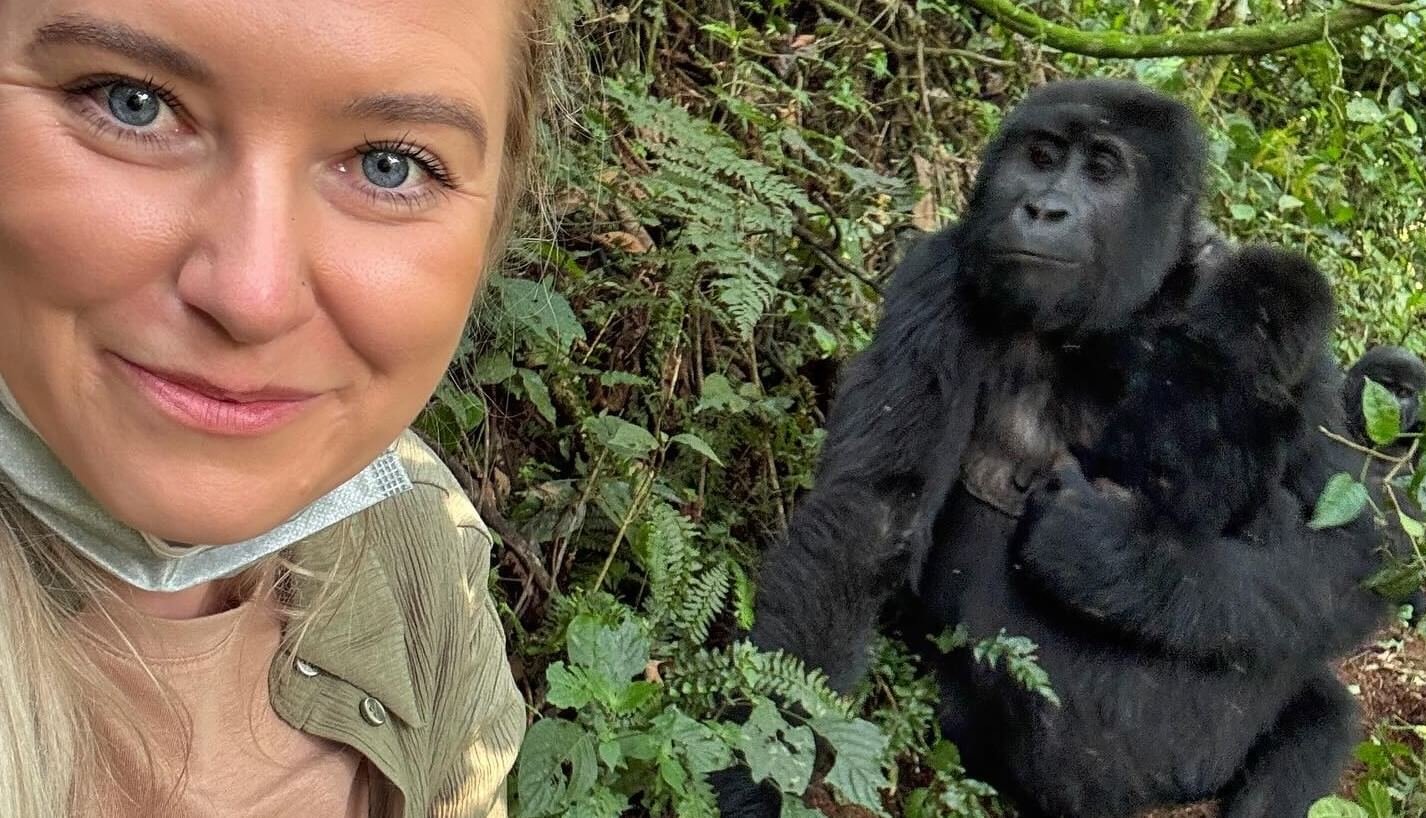 A close-up selfie of a female traveler with a large mountain gorilla visible just a few meters behind her in the forest undergrowth.