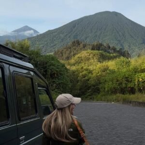 A traveler sitting by a safari vehicle, looking out at the lush green slopes of a volcanic mountain range.