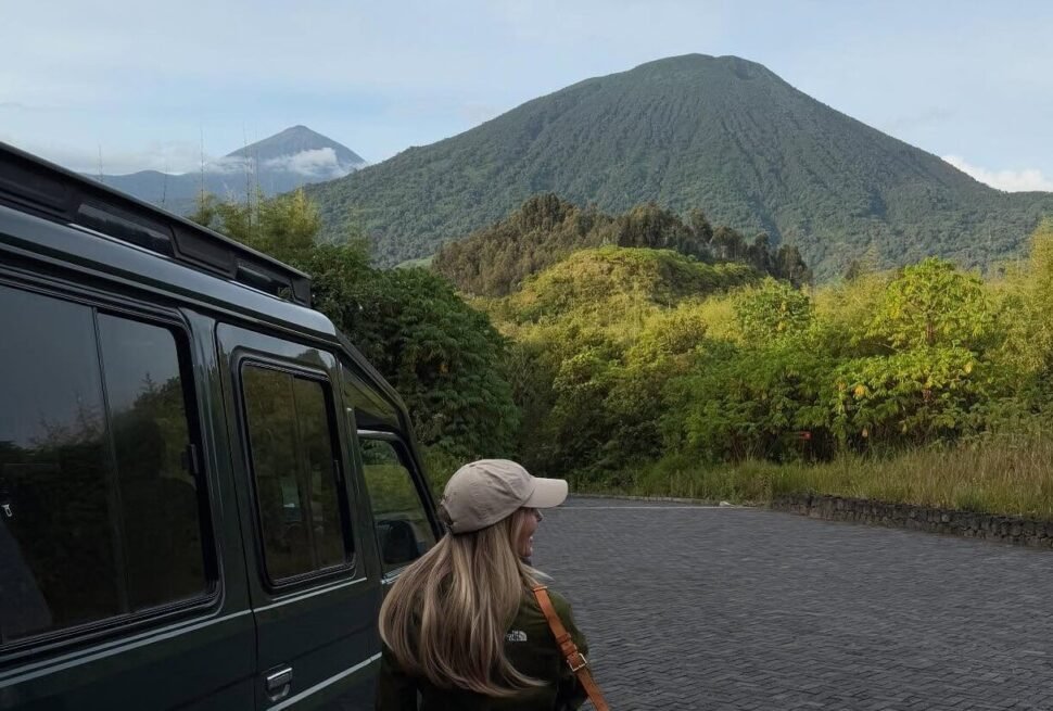 A traveler sitting by a safari vehicle, looking out at the lush green slopes of a volcanic mountain range.