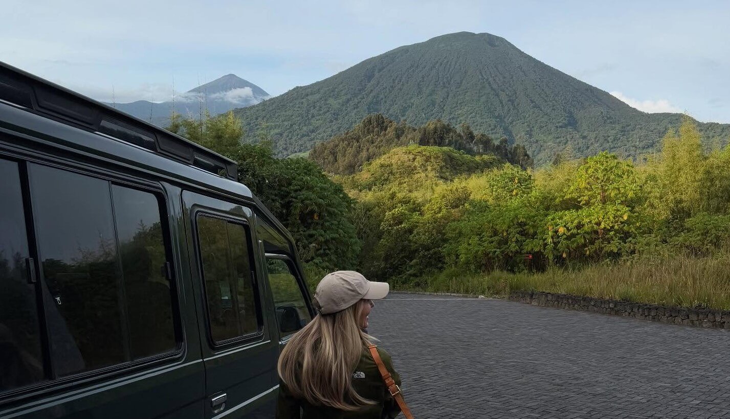 A traveler sitting by a safari vehicle, looking out at the lush green slopes of a volcanic mountain range.
