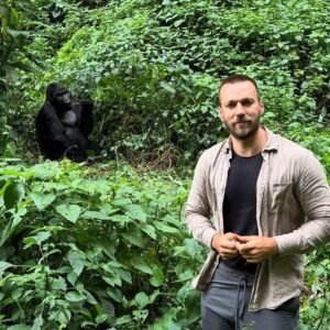 A male trekker standing on a narrow forest path with a silverback gorilla visible in the background, showing the proximity of the encounter.