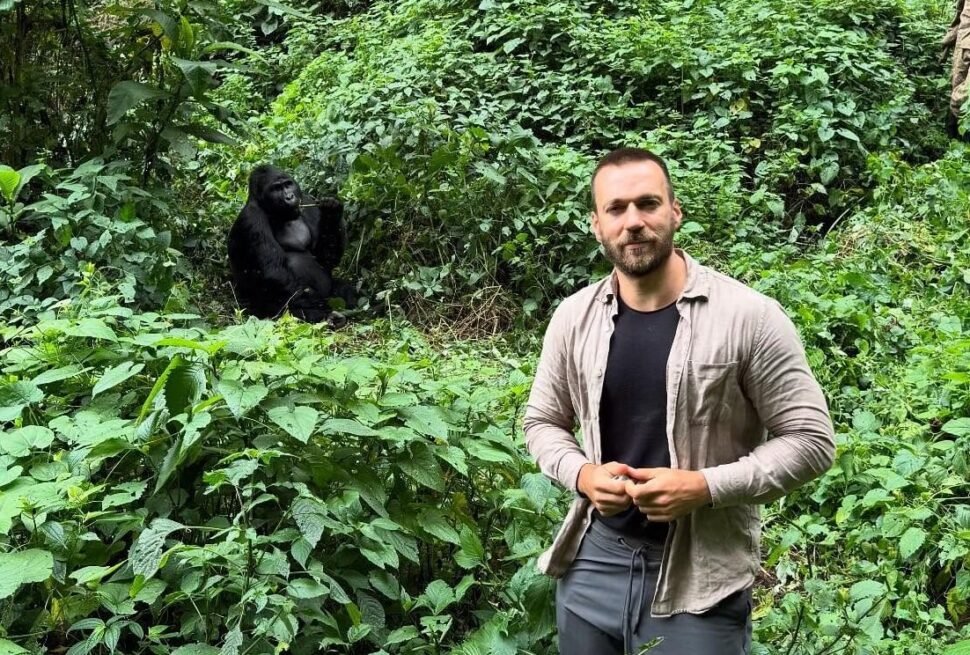 A male trekker standing on a narrow forest path with a silverback gorilla visible in the background, showing the proximity of the encounter.