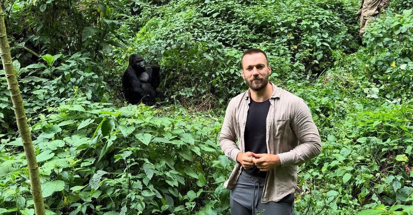 A male trekker standing on a narrow forest path with a silverback gorilla visible in the background, showing the proximity of the encounter.