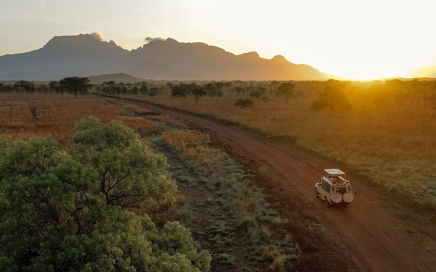 An aerial view of a safari vehicle driving along a dirt road through the misty hills of Bwindi at sunrise.